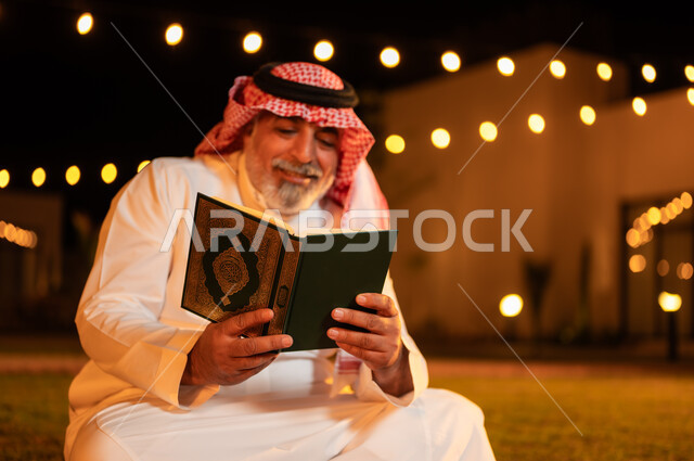 الجلوس في الهواء الطلق في حديقة شاليه فاخرة، التقرب الى الله في شهر رمضان المبارك، اداء الفرائض الدينية والاجتهاد في العبادة، تلاوة وحفظ المصحف الشريف في الاجواء الرمضانية الروحانية، رجل كبير بالسن عربي سعودي خليجي مسلم يرتدي الزي التقليدي يقرأ القرآن الكريم
