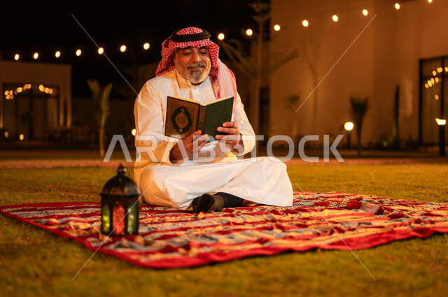 اداء الفرائض الدينية والاجتهاد في العبادة، تلاوة وحفظ المصحف الشريف في الاجواء الرمضانية الروحانية، رجل كبير بالسن عربي سعودي خليجي مسلم يرتدي الزي التقليدي يقرأ القرآن الكريم، الجلوس في الهواء الطلق في حديقة شاليه فاخرة، التقرب الى الله في شهر رمضان المبارك