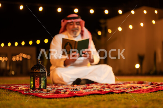 الاجتهاد في العبادة واداء الفرائض الدينية، تلاوة وحفظ المصحف الشريف في الاجواء الرمضانية الروحانية، رجل كبير بالسن عربي سعودي خليجي مسلم يرتدي الزي التقليدي يقرأ القرآن الكريم، الجلوس في الهواء الطلق في حديقة شاليه فاخرة، التقرب الى الله في شهر رمضان المبارك