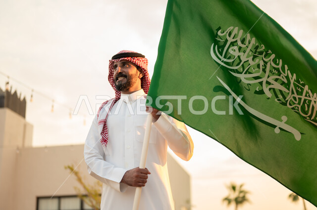 مفهوم حب الوطن والاعتزاز به, رجل عربي خليجي سعودي يرتدي الثوب التقليدي والشماغ يحمل بيده علم المملكة باحترام وفخر في حديقة المنزل, ذكرى يوم العلم 11مارس, استذكار اليوم الوطني السعودي في 23 سبتمبر, راية التوحيد والاسلام وشعار الهوية الوطنية