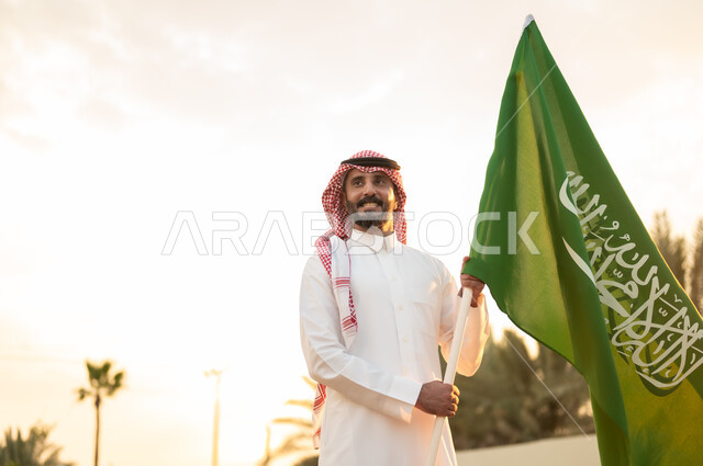 Commemoration of Flag Day on March 11, commemoration of the Saudi National Day on September 23, the banner of monotheism and Islam and the slogan of national identity, the concept of love for the homeland and pride in it, a Saudi Gulf Arab man wearing the traditional thobe and shemagh holding the Kingdom’s flag proudly in his hand.