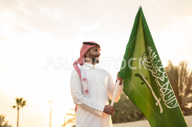  استذكار اليوم الوطني السعودي في 23 سبتمبر, راية التوحيد والاسلام وشعار الهوية الوطنية, مفهوم حب الوطن والاعتزاز به, رجل عربي خليجي سعودي يرتدي الثوب التقليدي والشماغ يحمل بيده علم المملكة باحترام, ذكرى يوم العلم 11مارس