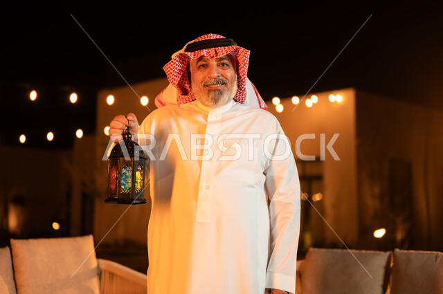 الأجواء الرمضانية الروحانية، الاحتفال بقدوم شهر رمضان المبارك، إضاء وزينة الأعياد والمناسبات في المملكة العربية السعودية, صورة مقربة لرجل كبير بالسن عربي خليجي سعودي يرتدي الشماغ والثوب التقليدي يمسك بيده فانوس معدني مضيء وينظر الى الكاميرا