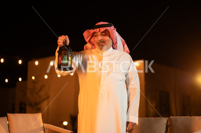 Lighting and decorations for holidays and events in the Kingdom of Saudi Arabia, a close-up of an elderly Saudi Gulf Arab man wearing a shemagh and traditional dress, holding a illuminated metal lantern in his hand and looking at it, the spiritual Ramadan atmosphere, happy family gatherings, celebrating the advent of the blessed month of Ramadan