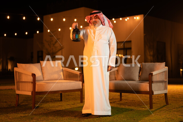 الاحتفال بقدوم شهر رمضان المبارك، إضاء وزينة الأعياد والمناسبات في المملكة العربية السعودية, رجل كبير بالسن عربي خليجي سعودي يرتدي الشماغ والثوب التقليدي يمسك بيده فانوس معدني مضيء وينظر إليه، الأجواء الرمضانية الروحانية، التجمعات العائلية السعيدة