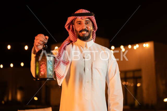 Lighting and decorations for holidays and events in the Kingdom of Saudi Arabia, a close-up image of a Saudi Gulf Arab man wearing a shemagh and traditional dress, holding a illuminated metal lantern in his hand and looking at the camera, the concept of family cohesion and happy family gatherings in the holy month of Ramadan