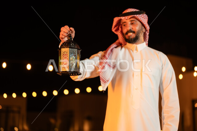 إضاء وزينة الأعياد والمناسبات في المملكة العربية السعودية, صورة مقربة لرجل عربي خليجي سعودي يرتدي الشماغ والثوب التقليدي يمسك بيده فانوس معدني مضيء وينظر إليه, مفهوم الترابط الأسري والجمعات العائلية السعيدة في شهر رمضان المبارك