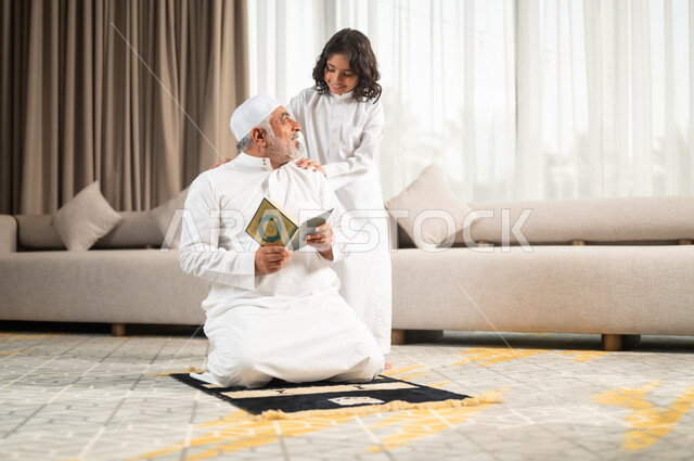 Diligent obedience and closeness to God, spiritual sessions in the blessed month of Ramadan, inculcating common sense in children, recitation and reading of the Holy Qur’an, an elderly Saudi Gulf Arab man wearing a white hat and traditional dress sitting on the prayer rug next to his grandson, performing religious duties.