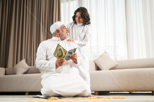 الجلسات الروحانية في شهر رمضان المبارك، زرع الفطرة السليمة في الاولاد، تلاوة وقراءة القرآن الكريم، رجل كبير عربي خليجي سعودي يرتدي الطاقية البيضاء والثوب التقليدي يجلس على سجادة الصلاة بجانب حفيده، اداء الفرائض الدينية، الاجتهاد بالطاعة والتقرب الى الله