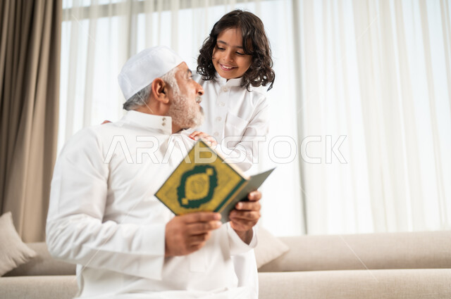 تلاوة وقراءة القرآن الكريم، رجل كبير عربي خليجي سعودي يرتدي الطاقية البيضاء والثوب التقليدي يجلس على سجادة الصلاة بجانب حفيده، اداء الفرائض الدينية، الاجتهاد بالطاعة والتقرب الى الله، الجلسات الروحانية في شهر رمضان المبارك، زرع الفطرة السليمة في الاولاد