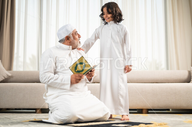 قراءة وتلاوة القرآن الكريم، رجل كبير عربي خليجي سعودي يرتدي الطاقية البيضاء والثوب التقليدي يجلس على سجادة الصلاة بجانب حفيده، اداء الفرائض الدينية، الاجتهاد بالطاعة والتقرب الى الله، الجلسات الروحانية في شهر رمضان المبارك، زرع الفطرة السليمة في الاولاد