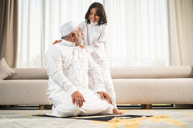 اداء الصلوات الخمس والفرائض الدينية، الاجتهاد بالطاعة والتقرب الى الله، الجلسات الروحانية في شهر رمضان المبارك، زرع الفطرة السليمة في الاولاد، رجل كبير عربي خليجي سعودي يرتدي الطاقية البيضاء والثوب التقليدي يجلس على سجادة الصلاة بجانب حفيده