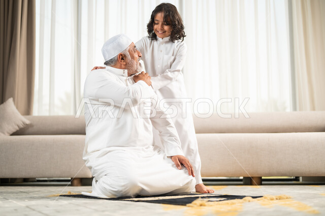  زرع الفطرة السليمة في الاولاد، رجل كبير عربي خليجي سعودي يرتدي الطاقية البيضاء والثوب التقليدي يجلس على سجادة الصلاة بجانب حفيده، اداء الصلوات الخمس والفرائض الدينية، الاجتهاد بالطاعة والتقرب الى الله، الجلسات الروحانية في شهر رمضان المبارك