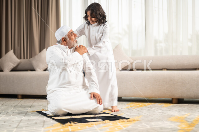اداء الصلوات الخمس والفرائض الدينية، الاجتهاد بالطاعة والتقرب الى الله، الجلسات الروحانية في شهر رمضان المبارك، زرع الفطرة السليمة في الاولاد، رجل كبير عربي خليجي سعودي يرتدي الطاقية البيضاء والثوب التقليدي يجلس على سجادة الصلاة بجانب حفيده