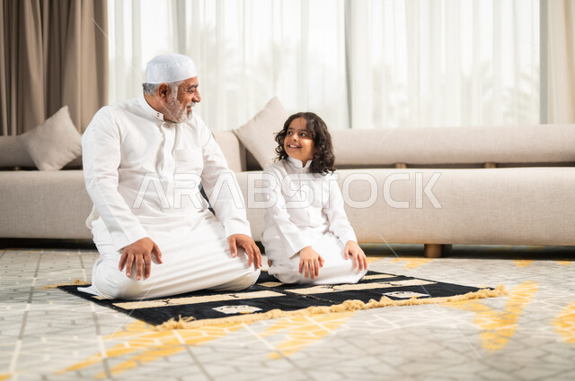  زرع الفطرة السليمة في الاولاد، رجل كبير عربي خليجي سعودي يرتدي الطاقية البيضاء والثوب التقليدي يجلس على سجادة الصلاة بجانب حفيده، اداء الصلوات الخمس والفرائض الدينية، الاجتهاد بالطاعة والتقرب الى الله، الجلسات الروحانية في شهر رمضان المبارك