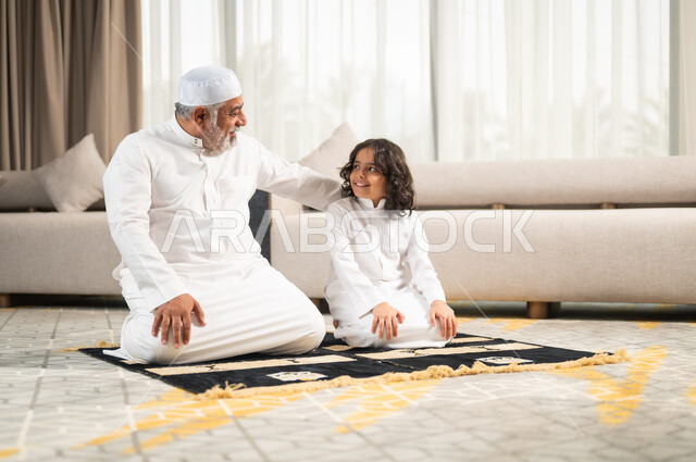  زرع الفطرة السليمة في الاولاد، رجل كبير عربي خليجي سعودي يرتدي الطاقية البيضاء والثوب التقليدي يجلس على سجادة الصلاة بجانب حفيده، اداء الصلوات الخمس والفرائض الدينية، الاجتهاد بالطاعة والتقرب الى الله، الجلسات الروحانية في شهر رمضان المبارك