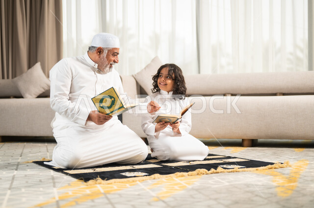 Diligent obedience and closeness to God, spiritual sessions in the blessed month of Ramadan, inculcating good sense in children, reading and reciting the Holy Qur’an, an elderly Saudi Gulf Arab man wearing a white hat and traditional dress sitting on the prayer rug next to his grandson, performing religious duties.