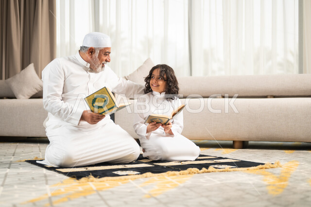 Reading and reciting the Holy Qur’an, an elderly Saudi Gulf Arab man wearing a white hat and traditional dress sitting on the prayer rug next to his grandson, performing religious duties, striving to obey and get closer to God, spiritual sessions in the blessed month of Ramadan, instilling common sense in children.
