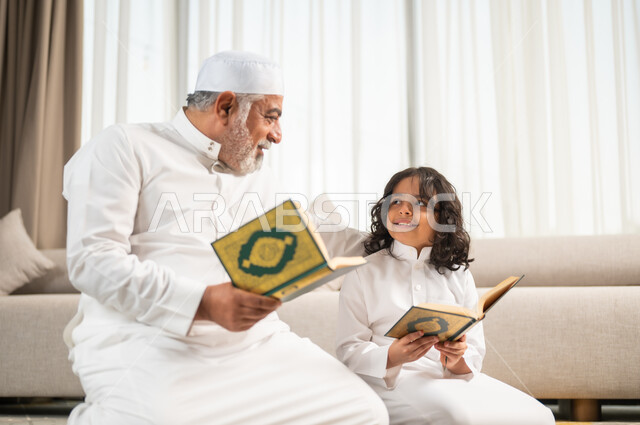 الجلسات الروحانية في شهر رمضان المبارك، زرع الفطرة السليمة في الاولاد، قراءة وتلاوة القرآن الكريم، رجل كبير عربي خليجي سعودي يرتدي الطاقية البيضاء والثوب التقليدي يجلس على سجادة الصلاة بجانب حفيده، اداء الفرائض الدينية، الاجتهاد بالطاعة والتقرب الى الله
