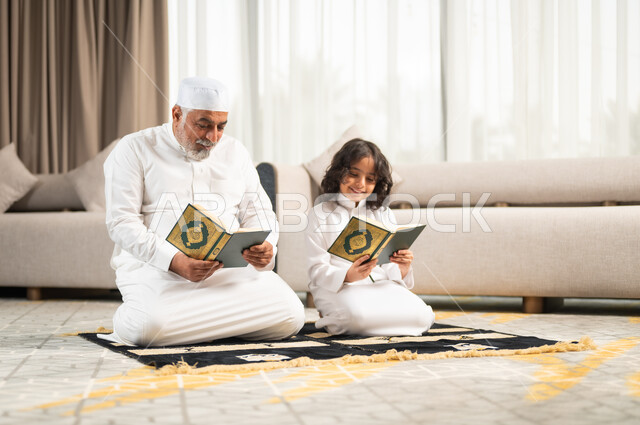 قراءة وتلاوة القرآن الكريم، رجل كبير عربي خليجي سعودي يرتدي الطاقية البيضاء والثوب التقليدي يجلس على سجادة الصلاة بجانب حفيده، اداء الفرائض الدينية، الاجتهاد بالطاعة والتقرب الى الله، الجلسات الروحانية في شهر رمضان المبارك، زرع الفطرة السليمة في الاولاد