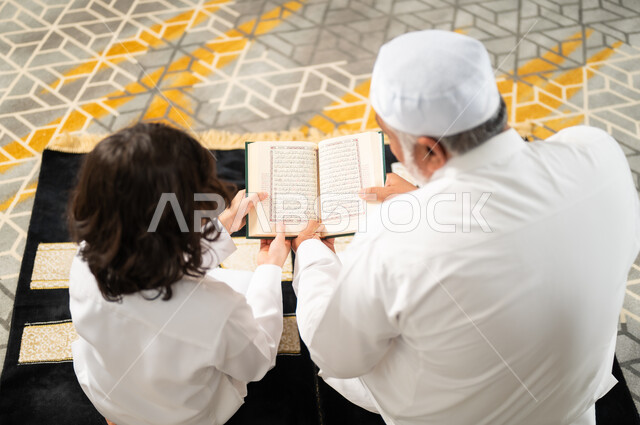 الجلسات الروحانية في شهر رمضان، زرع الفطرة السليمة في الاولاد، قراءة القرآن الكريم، صورة من الخلف لرجل كبير عربي خليجي سعودي يرتدي الطاقية البيضاء والثوب التقليدي يجلس على سجادة الصلاة بجانب حفيده، اداء الفرائض الدينية، الاجتهاد بالطاعة والتقرب الى الله