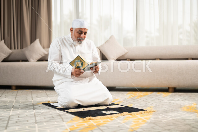  تلاوة وقراءة القرآن الكريم، الإكثار من الطاعة وذكر الله تعالى، رجل كبير بالسن عربي سعودي خليجي مسلم يجلس على سجادة الصلاة في غرفة المعيشة يمسك بيده المصحف الشريف، التقرب الى رب العالمين في شهر رمضان المبارك، الاجتهاد في العبادة واداء الفرائض الدينية