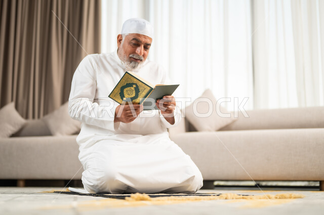 قراءة و تلاوة القرآن الكريم، الإكثار من الطاعة وذكر الله تعالى، رجل كبير بالسن عربي سعودي خليجي مسلم يجلس على سجادة الصلاة في غرفة المعيشة يمسك بيده المصحف الشريف، التقرب الى رب العالمين في شهر رمضان المبارك، الاجتهاد في العبادة واداء الفرائض الدينية