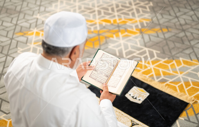 الإكثار من الطاعة وذكر الله تعالى، الاجتهاد في العبادة واداء الفرائض الدينية، قراءة و تلاوة القرآن الكريم في شهر رمضان المبارك، صورة من الخلف لرجل كبير بالسن عربي سعودي خليجي مسلم يجلس على سجادة الصلاة في غرفة المعيشة يمسك بيده المصحف الشريف