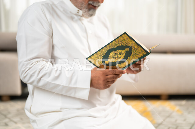  الاجتهاد في العبادة واداء الفرائض الدينية، قراءة و تلاوة القرآن الكريم، الإكثار من الطاعة وذكر الله تعالى، رجل كبير بالسن عربي سعودي خليجي مسلم يجلس على سجادة الصلاة في غرفة المعيشة يمسك بيده المصحف الشريف، التقرب الى رب العالمين في شهر رمضان المبارك