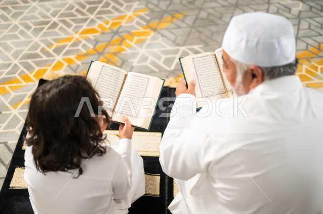 الجلسات الروحانية في شهر رمضان، زرع الفطرة السليمة في الاولاد، قراءة القرآن الكريم، صورة من الخلف لرجل كبير عربي خليجي سعودي يرتدي الطاقية البيضاء والثوب التقليدي يجلس على سجادة الصلاة بجانب حفيده، اداء الفرائض الدينية، الاجتهاد بالطاعة والتقرب الى الله