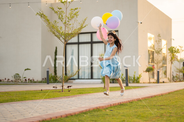 إظهار مشاعر السرور والفرح بقدوم العيد, الاحتفال في الأعياد والمناسبات في الهواء الطلق, فتاة عربية خليجية سعودية ترتدي الفستان الأبيض تمسك بيدها مجموعة من البلالين الملونة تركض في حديقة المنزل, أجواء العيد في المملكة العربية السعودية