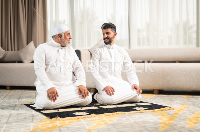 الإكثار من الطاعات وذكر الله تعالى، رجل كبير بالسن عربي سعودي خليجي يجلس على سجادة الصلاة في غرفة المعيشة يؤدي فرض الصلاة بجانب ابنه الشاب، التقرب الى رب العالمين في شهر رمضان المبارك، الاجتهاد في العبادة واداء الفرائض الدينية، اجواء عائلية رمضانية روحانية
