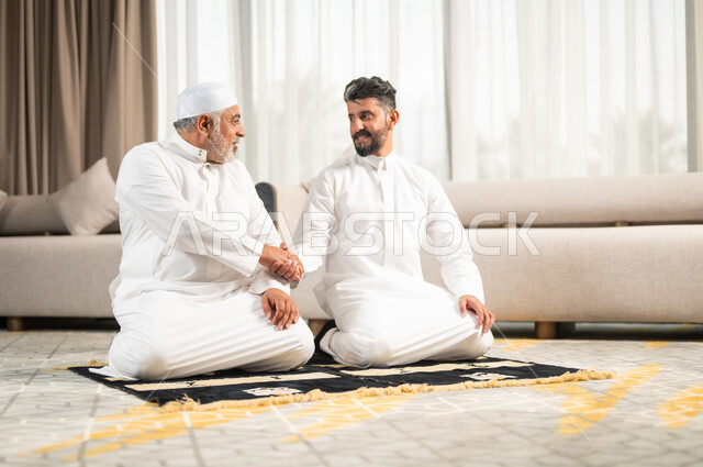 التقرب الى رب العالمين في شهر رمضان المبارك، الاجتهاد في العبادة واداء الفرائض الدينية، اجواء عائلية رمضانية روحانية، الإكثار من الطاعات وذكر الله تعالى، رجل كبير بالسن عربي سعودي خليجي يجلس على سجادة الصلاة في غرفة المعيشة يؤدي فرض الصلاة بجانب ابنه الشاب