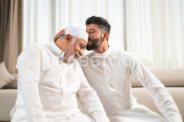 الاجتهاد في العبادة واداء الفرائض الدينية، اجواء عائلية رمضانية روحانية، الإكثار من الطاعات وذكر الله تعالى، شاب عربي سعودي خليجي يجلس على سجادة الصلاة في غرفة المعيشة يؤدي فرض الصلاة يقوم بتقبيل رأس والده، التقرب الى رب العالمين في شهر رمضان المبارك