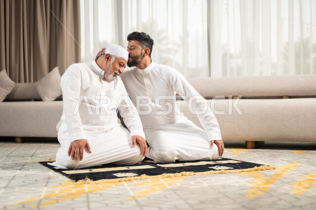 A spiritual Ramadan family atmosphere, increasing obedience and remembrance of God Almighty, a young Saudi Arab Gulf man sitting on the prayer rug in the living room performing the obligatory prayer, kissing his father’s head, getting closer to the Lord of the Worlds in the blessed month of Ramadan, diligent in worship and performing religious obligations.