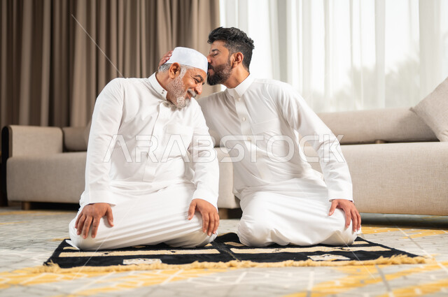 الاجتهاد في العبادة واداء الفرائض الدينية، اجواء عائلية رمضانية روحانية، الإكثار من الطاعات وذكر الله تعالى، شاب عربي سعودي خليجي يجلس على سجادة الصلاة في غرفة المعيشة يؤدي فرض الصلاة يقوم بتقبيل رأس والده، التقرب الى رب العالمين في شهر رمضان المبارك