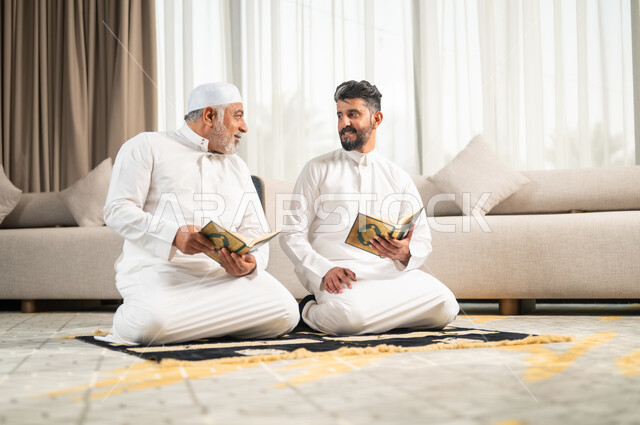  اجواء عائلية رمضانية روحانية، الإكثار من الطاعات وذكر الله تعالى، شاب عربي سعودي خليجي يجلس على سجادة الصلاة في غرفة المعيشة بجانب والده يقومان بقراءة القرآن الكريم، تلاوة وحفظ المصحف الشريف في شهر رمضان المبارك، الاجتهاد في العبادة واداء الفرائض الدينية