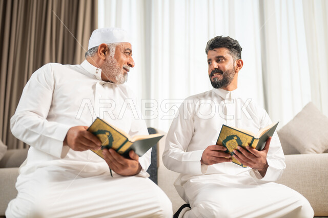 تلاوة وحفظ المصحف الشريف في شهر رمضان المبارك، الاجتهاد في العبادة واداء الفرائض الدينية، اجواء عائلية رمضانية روحانية، الإكثار من الطاعات وذكر الله تعالى، شاب عربي سعودي خليجي يجلس على سجادة الصلاة في غرفة المعيشة بجانب والده يقومان بقراءة القرآن الكريم