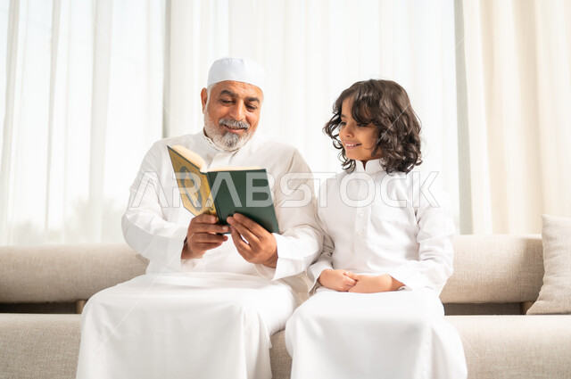 الجلسات الروحانية في شهر رمضان المبارك، زرع الفطرة السليمة في الاولاد، رجل كبير عربي خليجي سعودي يرتدي الطاقية البيضاء والثوب التقليدي يجلس على الاريكة بجانب حفيده يقومان  بقراءة وتلاوة القرآن الكريم، اداء الفرائض الدينية، الاجتهاد بالطاعة والتقرب الى الله