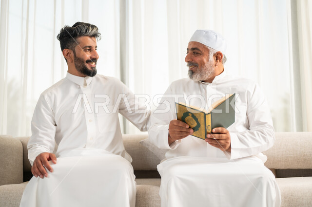  اجواء عائلية رمضانية روحانية، الإكثار من الطاعات وذكر الله تعالى، شاب عربي سعودي خليجي يجلس على الاريكة في غرفة المعيشة بجانب والده يقومان بقراءة القرآن الكريم، تلاوة وحفظ المصحف الشريف في شهر رمضان المبارك، الاجتهاد في العبادة واداء الفرائض الدينية