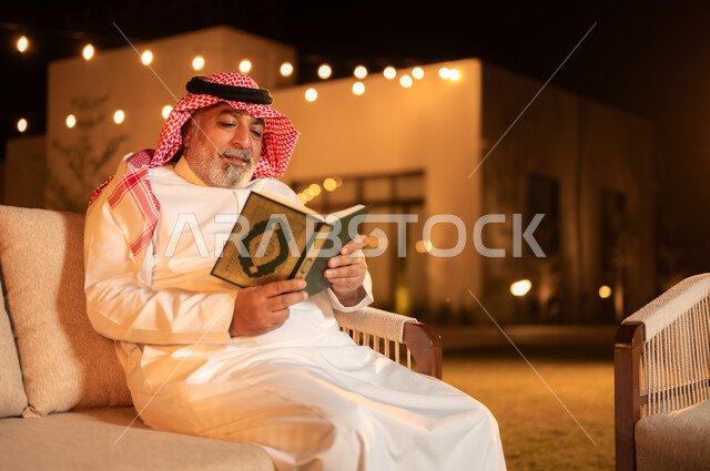 تلاوة وحفظ المصحف الشريف، رجل كبير بالسن عربي سعودي خليجي مسلم يرتدي الشماغ والثوب التقليدي يقوم بقراءة القرآن الكريم، الجلوس في الهواء الطلق في حديقة احدى الشاليهات الفاخرة، التقرب الى الله في شهر رمضان المبارك, الاجتهاد في العبادة واداء الفرائض الدينية