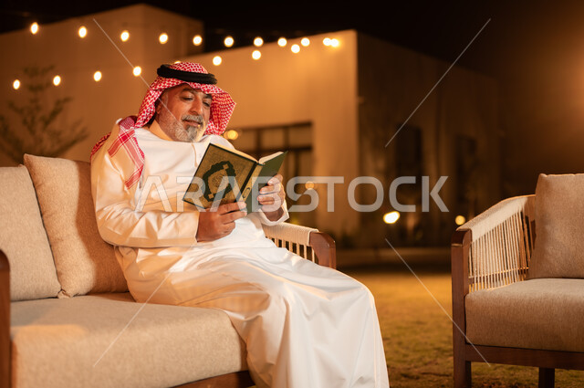التقرب الى الله في شهر رمضان المبارك, الاجتهاد في العبادة واداء الفرائض الدينية، تلاوة وحفظ المصحف الشريف، رجل كبير بالسن عربي سعودي خليجي مسلم يرتدي الشماغ والثوب التقليدي يقوم بقراءة القرآن الكريم، الجلوس في الهواء الطلق في حديقة احدى الشاليهات الفاخرة