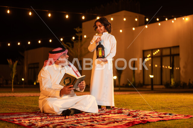 الإكثار من الطاعة والتقرب الى الله، الجلسات الإسلامية الخارجية الهادئة في شهر رمضان المبارك، زرع الفطرة السليمة الصحيحة في الأبناء، تلاوة وقراءة القرآن الكريم، رجل عربي خليجي سعودي كبير بالسن يحمل بيده المصحف الشريف يجلس على العشب مع حفيده في إحدى الشاليهات