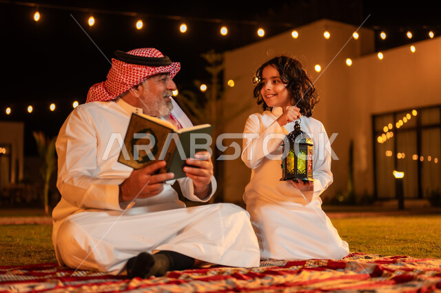 Cultivating the correct common sense in children, reciting and reading the Holy Qur’an, increasing obedience and drawing closer to God, quiet outdoor Islamic sessions in the blessed month of Ramadan, an elderly Saudi Arabian Gulf man holding the Holy Qur’an in his hand sitting on the grass with his grandson in one of the chalets.