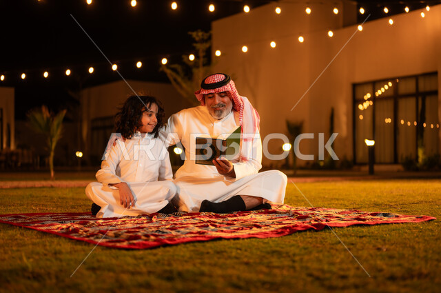 الإكثار من الطاعة والتقرب الى الله، الجلسات الإسلامية الخارجية الهادئة في شهر رمضان المبارك، زرع الفطرة السليمة الصحيحة في الأبناء، تلاوة وقراءة القرآن الكريم، رجل عربي خليجي سعودي كبير بالسن يحمل بيده المصحف الشريف يجلس على العشب مع حفيده في إحدى الشاليهات
