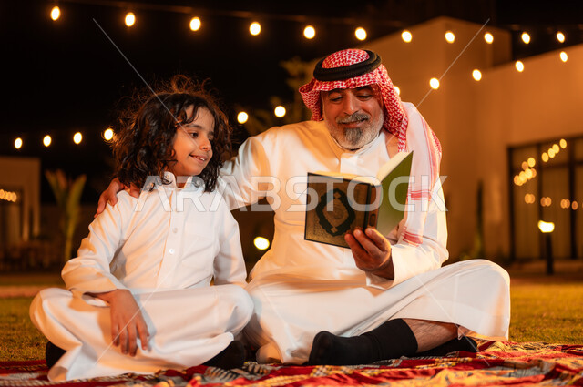زرع الفطرة السليمة الصحيحة في الأبناء، تلاوة وقراءة القرآن الكريم، الإكثار من الطاعة والتقرب الى الله، الجلسات الإسلامية الخارجية الهادئة في شهر رمضان المبارك، رجل عربي خليجي سعودي كبير بالسن يحمل بيده المصحف الشريف يجلس على العشب مع حفيده في إحدى الشاليهات
