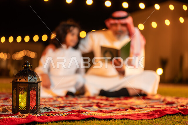 تلاوة وقراءة القرآن الكريم، الإكثار من الطاعة والتقرب الى الله، الجلسات الإسلامية الخارجية الهادئة في شهر رمضان المبارك، زرع الفطرة السليمة الصحيحة في الأبناء، رجل عربي خليجي سعودي كبير بالسن يحمل بيده المصحف الشريف يجلس على العشب مع حفيده في إحدى الشاليهات