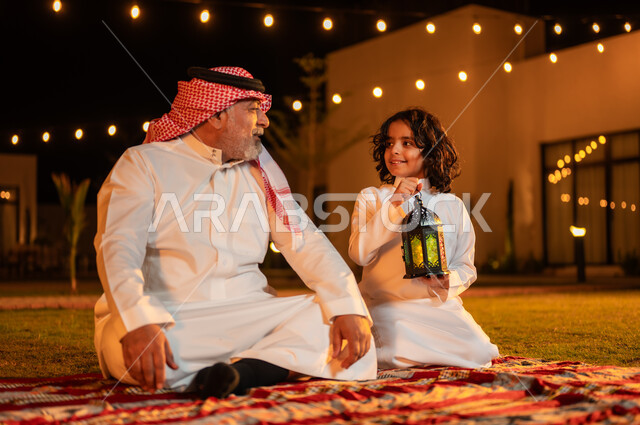  الجلسات الخارجية الهادئة في شهر رمضان المبارك، قضاء أوقات عائلية ممتعة في إحدى الشاليهات، تعليم الأبناء العادات والتقاليد الإسلامية، ولد عربي خليجي سعودي يرتدي الثوب التقليدي يجلس مع جده يحمل بيده فانوس معدني مزخرف مضيء