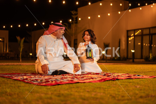  الجلسات الخارجية الهادئة في شهر رمضان المبارك، قضاء أوقات عائلية ممتعة في إحدى الشاليهات، تعليم الأبناء العادات والتقاليد الإسلامية، ولد عربي خليجي سعودي يرتدي الثوب التقليدي يجلس مع جده يحمل بيده فانوس معدني مزخرف مضيء