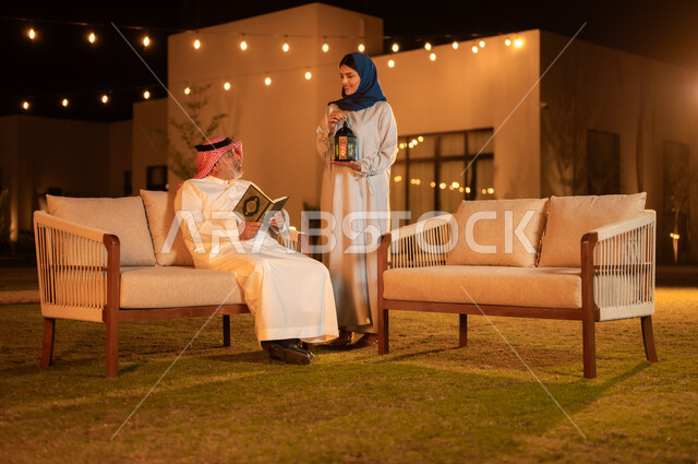 التقرب إلى الله في شهر رمضان المبارك, تلاوة وحفظ المصحف الشريف، رجل كبير بالسن عربي سعودي خليجي يرتدي الشماغ والثوب التقليدي يحمل بيده القرآن الكريم، الجلوس في الهواء الطلق في حديقة إحدى الشاليهات الفاخرة، امرأة محجبة تحمل بيدها فانوس معدني مزخرف مضيء